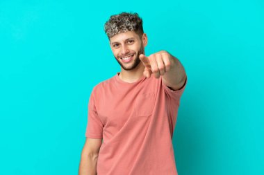 Mavi arka planda izole edilmiş yakışıklı beyaz adam mutlu bir ifadeyle ön tarafı işaret ediyor.