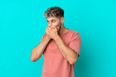 Genç beyaz adam mavi arka planda izole edilmiş ağzını kapatıyor ve yan tarafa bakıyor.