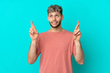 Mavi arka planda izole edilmiş genç, yakışıklı beyaz bir adam parmaklarını çapraz yapıp en iyisini diliyor.