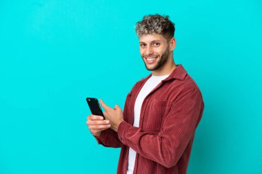 Young handsome caucasian man isolated on blue background using mobile phone and pointing back