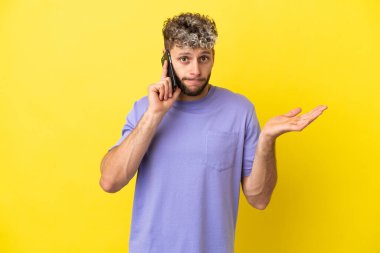 Sarı arka planda izole edilmiş cep telefonu kullanan genç beyaz adam ellerini kaldırırken şüpheye düşüyor.