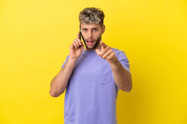 Sarı arka planda izole edilmiş cep telefonu kullanan genç beyaz adam şaşırmış ve ön tarafı işaret ediyor.