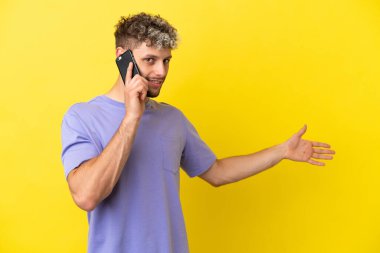 Young caucasian man using mobile phone isolated on yellow background extending hands to the side for inviting to come