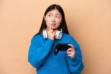 Young Chinese man playing with a video game controller isolated on beige background having doubts while looking up