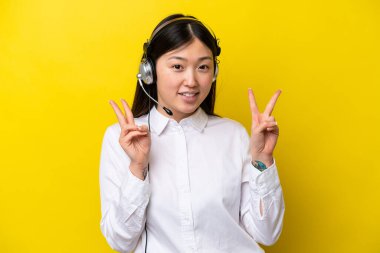 Telefon pazarlamacısı Çinli bir kadın sarı arka planda izole edilmiş bir kulaklıkla çalışıyor. İki eliyle zafer işareti gösteriyor.