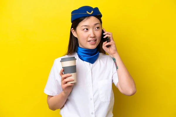 Airplane Chinese woman stewardess isolated on yellow background holding coffee to take away and a mobile