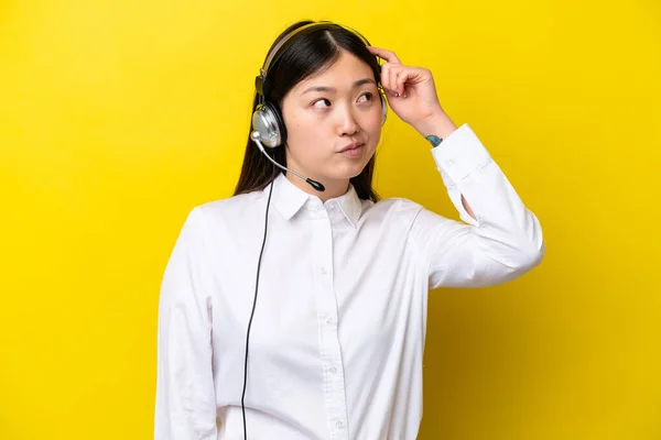 Telemarketer Chinese woman working with a headset isolated on yellow background having doubts and with confuse face expression