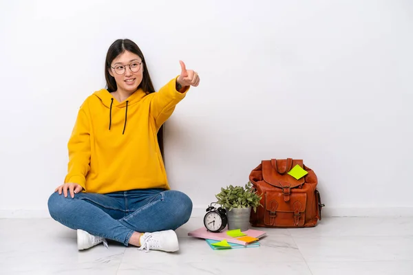 stock image Young Chinese student woman sitting on the floor isolated on white wall giving a thumbs up gesture