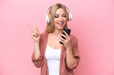Pretty blonde woman isolated on pink background listening music with a mobile and singing