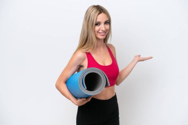Genç sarışın kadın yoga derslerine giderken beyaz arka planda izole edilmiş bir minder tutuyor ve davet etmek için ellerini yana uzatıyor.