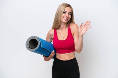 Genç sporcu sarışın kadın yoga derslerine giderken beyaz arka planda izole edilmiş bir minder tutuyor ve mutlu bir ifadeyle selam veriyor.