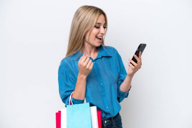 Güzel sarışın kadın beyaz arka planda izole edilmiş alışveriş torbaları tutuyor ve bir arkadaşına cep telefonuyla mesaj yazıyor.