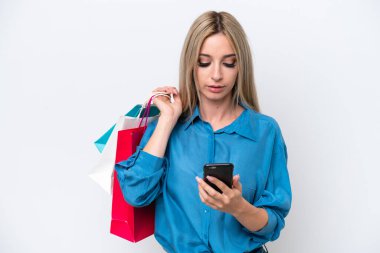 Pretty blonde woman isolated on white background holding shopping bags and writing a message with her cell phone to a friend