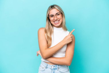 Mavi arka planda izole edilmiş genç beyaz kadın bir ürünü sunmak için kenarı işaret ediyor.