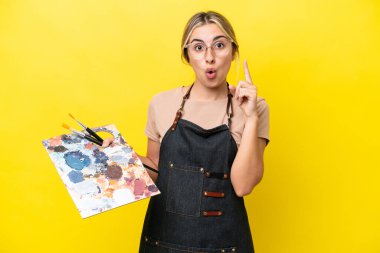 Young artist caucasian woman holding a palette isolated  on yellow background thinking an idea pointing the finger up