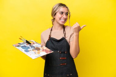 Sarı arka planda izole edilmiş palet tutan beyaz bir kadın bir ürünü sunmak için yan tarafı işaret ediyor.