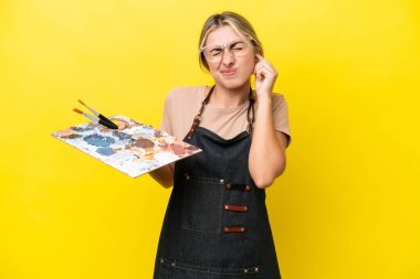 Young artist caucasian woman holding a palette isolated  on yellow background frustrated and covering ears