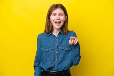 Sarı arka planda izole edilmiş genç bir İngiliz kadın şaşırmış ve ön tarafı işaret ediyor.