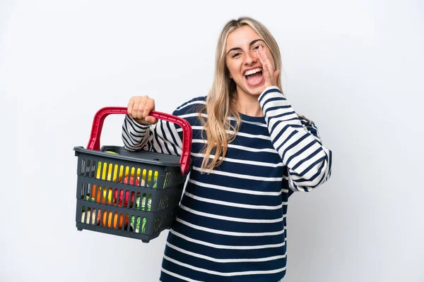 Young caucasian woman holding a shopping basket full of food isolated on white background shouting with mouth wide open