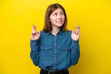 Sarı arka planda izole edilmiş genç bir İngiliz kadın parmaklarını çapraz yapıp en iyisini diliyor.