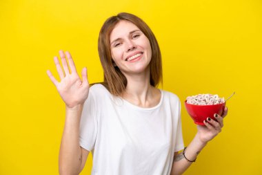 Sarı arka planda izole edilmiş kahve bardağını tutan genç İngiliz kadın mutlu bir ifadeyle selamlıyor.