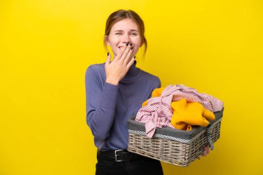 Sarı arka planda izole edilmiş çamaşır sepetini tutan genç İngiliz kadın mutlu ve ağzı kapalı gülümsüyor.