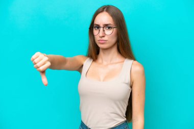 Mavi arka planda izole edilmiş genç beyaz kadın olumsuz ifadeyle baş parmağını aşağı gösteriyor.