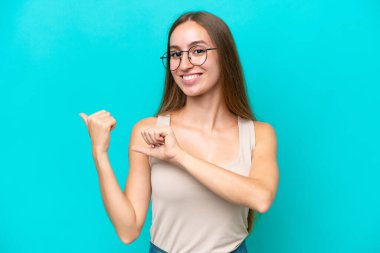 Mavi arka planda izole edilmiş genç beyaz kadın bir ürünü sunmak için kenarı işaret ediyor.