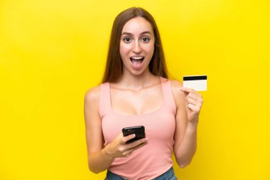 Sarı arka planda izole edilmiş genç beyaz bir kadın cep telefonuyla alışveriş yapıyor ve elinde şaşırmış bir ifadeyle bir kredi kartı tutuyor.