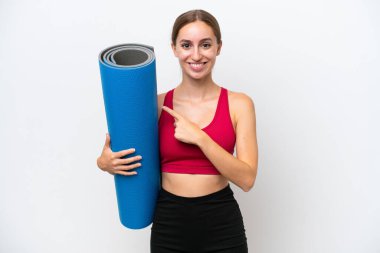 Beyaz arka planda izole edilmiş bir minder tutarak yoga derslerine giden beyaz bir kadın bir ürünü sunmak için yan tarafı işaret ediyor.