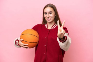 Kafkasyalı genç basketbolcu kadın pembe arka planda izole edilmiş gülümsüyor ve zafer işareti gösteriyor.