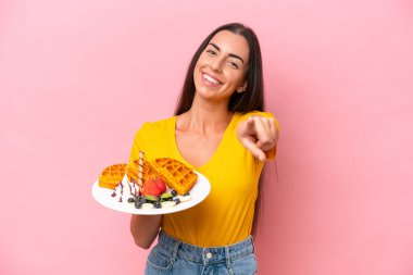 Pembe arka planda gözleme tutan genç beyaz kadın mutlu bir ifadeyle ön tarafı işaret ediyor.