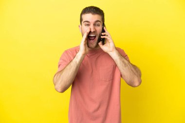 Yakışıklı sarışın adam izole edilmiş arka planda cep telefonu kullanıyor ve ağzı açık bağırıyor.