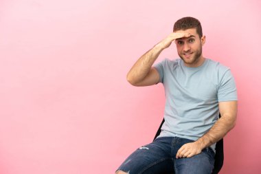 İzole edilmiş pembe arka planda bir sandalyede oturan genç adam bir şeye bakmak için uzaklara bakıyor.