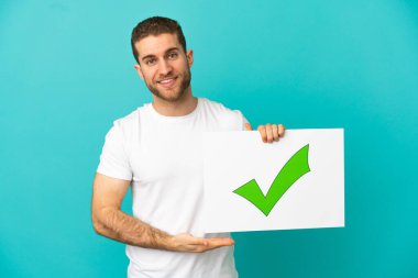 İzole edilmiş mavi arkaplan üzerinde yakışıklı sarışın adam içinde yeşil işaretli ve mutlu ifadeli bir pankart tutuyor.
