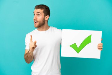 Yakışıklı sarışın adam izole edilmiş mavi arka planda elinde yeşil işaret simgesi olan bir pankart tutuyor ve düşünüyor.