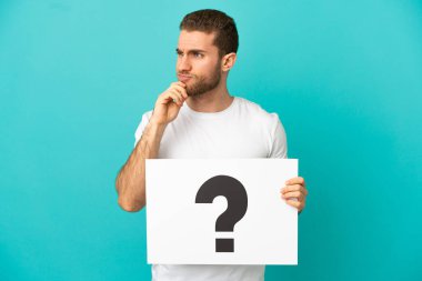 İzole edilmiş mavi arka planda yakışıklı sarışın bir adam soru işareti olan bir pankart tutuyor ve düşünüyor.