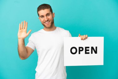 Yakışıklı, mavi arkaplan üzerinde açık bir pankart tutan ve selam veren sarışın bir adam.