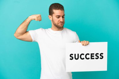 Yakışıklı sarışın, mavi arka planda. Elinde yazılı bir levha var ve güçlü bir jest yapıyor.