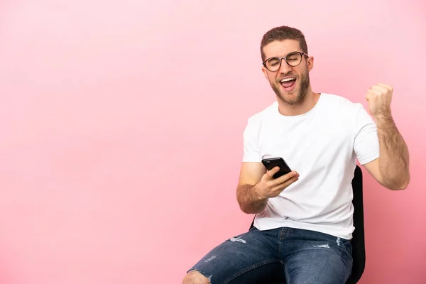Young Man Sitting Chair Isolated Pink Background Phone Victory Position — Stock Photo, Image