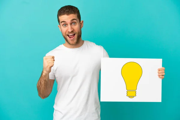 Handsome Blonde Man Isolated Blue Background Holding Placard Bulb Icon — Stock fotografie