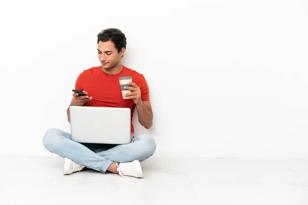 Caucasian handsome man with a laptop sitting on the floor holding coffee to take away and a mobile