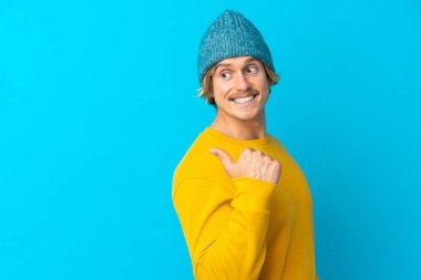 Mavi arka planda izole edilmiş yakışıklı sarışın bir adam bir ürün sunmak için kenarı işaret ediyor.