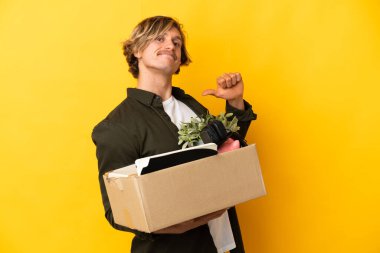 Sarı arka planda izole edilmiş, gururlu ve kendini beğenmiş şeylerle dolu bir kutuyu kaldırırken sarışın adam hamle yapıyor.
