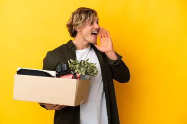 Sarı arka planda izole edilmiş şeylerle dolu bir kutuyu kaldırırken sarışın adam hareket ediyor ve ağzı sonuna kadar açık bağırıyor.