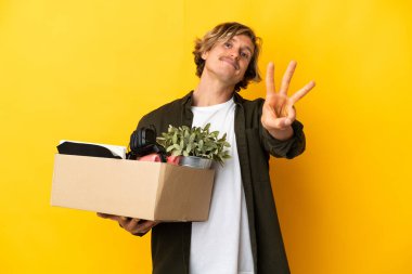 Sarışın adam, sarı arka planda izole edilmiş şeylerle dolu bir kutuyu kaldırırken hareket ediyor mutlu ve parmakları olan üç kişi sayıyor.
