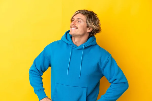 Handsome blonde man isolated on yellow background posing with arms at hip and smiling