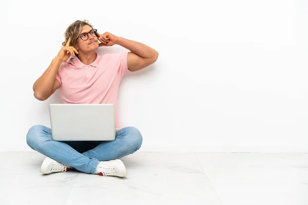 Young caucasian man sitting on the floor with his laptop isolated on white background having doubts and thinking