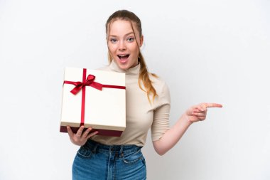 Beyaz arka planda izole edilmiş bir hediye tutan genç beyaz kadın şaşırdı ve parmağını yana çevirdi.
