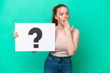 Young caucasian woman isolated on green background holding a placard with question mark symbol with surprised expression